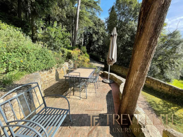 Уютная терраса на природе с деревянным столом, металлическими стульями и зонтом, окруженная зеленью и деревьями, идеально подходит для отдыха и приятных встреч на свежем воздухе.