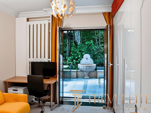 Современная квартира с выходом на террасу, интерьер с яркой мебелью и панорамными окнами, создающими уют и простор, а за стеклом — зеленый сад и декоративный фонтан.