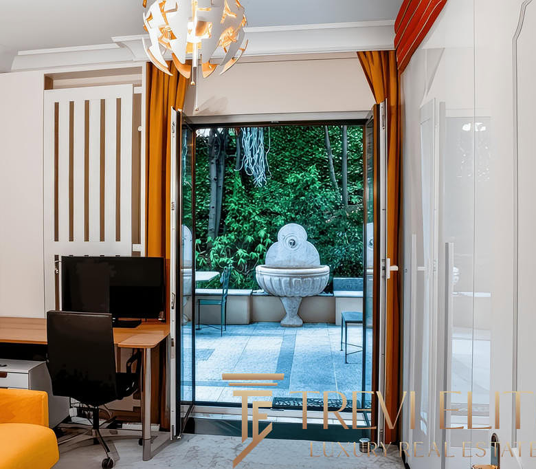 Современная квартира с выходом на террасу, интерьер с яркой мебелью и панорамными окнами, создающими уют и простор, а за стеклом — зеленый сад и декоративный фонтан.