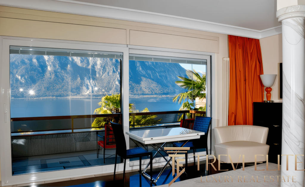 Вид из квартиры на живописное озеро и горные пейзажи через большие панорамные окна. Современный интерьер с уютной зоной отдыха и обеденной зоной, яркие акценты в декоре, создающие атмосферу комфорта и роскоши.