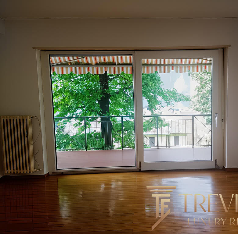 Окно с видом на зеленый парк и балкон через стеклянные двери, интерьер с деревянным полом и светлыми стенами, агентство недвижимости Треви Элит.
