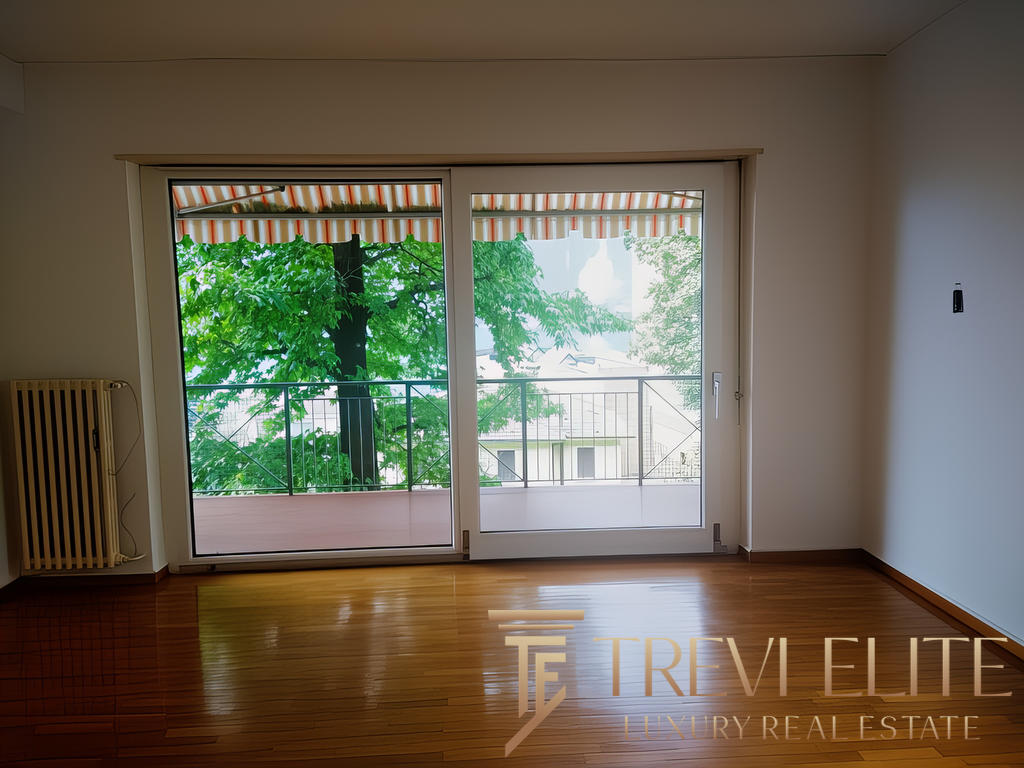 Окно с видом на зеленый парк и балкон через стеклянные двери, интерьер с деревянным полом и светлыми стенами, агентство недвижимости Треви Элит.