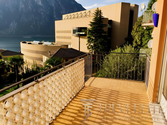 вид с балкона на современное здание у озера и горный пейзаж, солнечная погода, зелень, комфортное пространство для отдыха и наслаждения природой и видами.