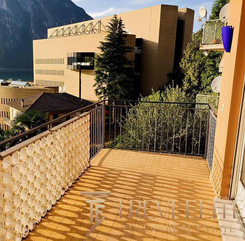 вид с балкона на современное здание у озера и горный пейзаж, солнечная погода, зелень, комфортное пространство для отдыха и наслаждения природой и видами.