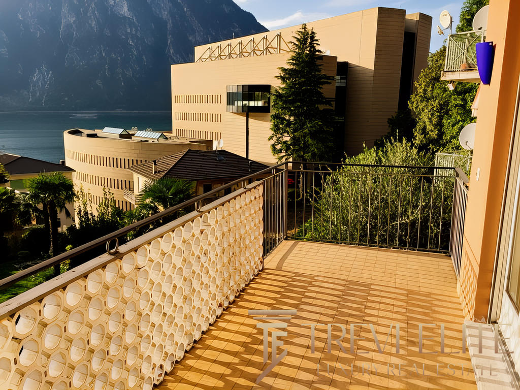 вид с балкона на современное здание у озера и горный пейзаж, солнечная погода, зелень, комфортное пространство для отдыха и наслаждения природой и видами.
