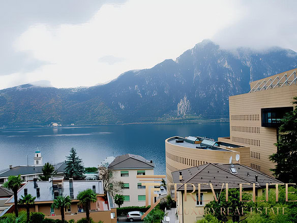 Красивая панорамная фотография горного озера и современных жилых домов на берегу, окружённых зеленью, с видом на покрытые облаками вершины.