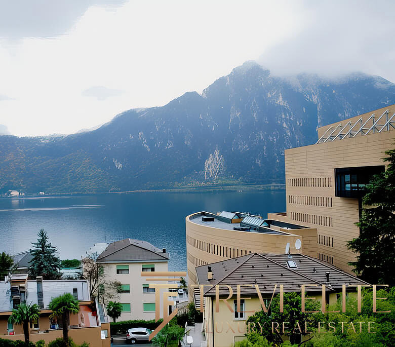 Красивая панорамная фотография горного озера и современных жилых домов на берегу, окружённых зеленью, с видом на покрытые облаками вершины.