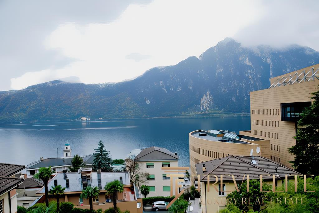 Красивая панорамная фотография горного озера и современных жилых домов на берегу, окружённых зеленью, с видом на покрытые облаками вершины.