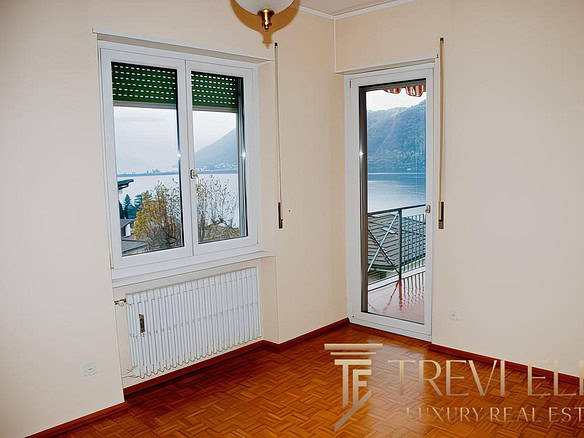 Вид из окна в квартире с балконом, панорамный взгляд на озеро и горы, светлые стены и деревянный пол, уютная обстановка для комфортного жилья.