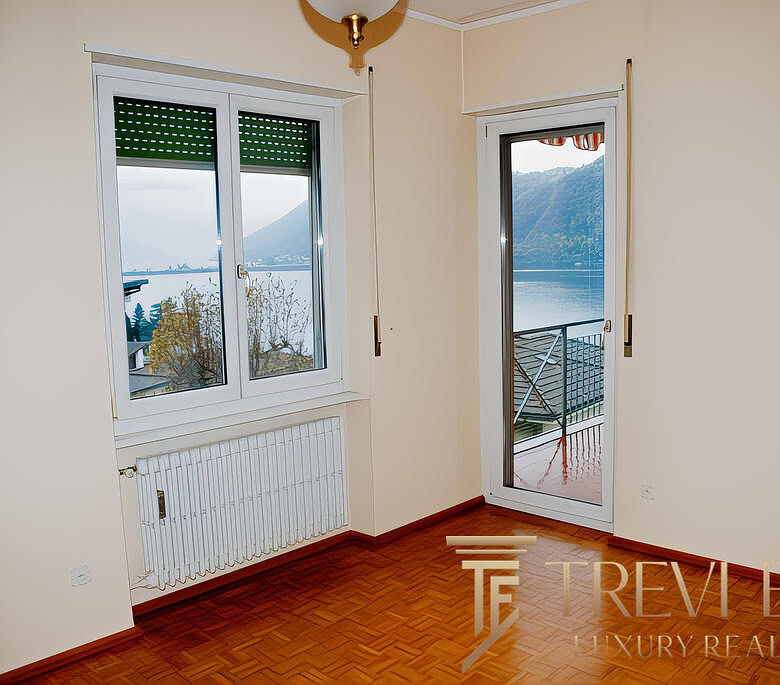 Вид из окна в квартире с балконом, панорамный взгляд на озеро и горы, светлые стены и деревянный пол, уютная обстановка для комфортного жилья.