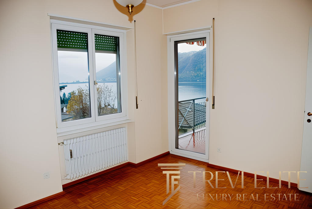 Вид из окна в квартире с балконом, панорамный взгляд на озеро и горы, светлые стены и деревянный пол, уютная обстановка для комфортного жилья.