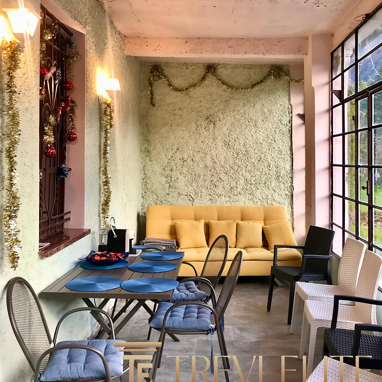 Уютная зона для отдыха и общения с декоративными елками и гирляндами, большие панорамные окна, мягкий диван и стулья, праздничное оформление интерьера в светлых тонах.