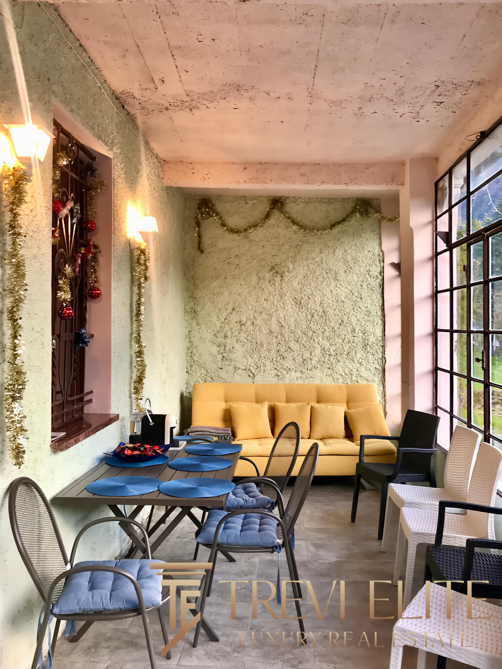 Уютная зона для отдыха и общения с декоративными елками и гирляндами, большие панорамные окна, мягкий диван и стулья, праздничное оформление интерьера в светлых тонах.