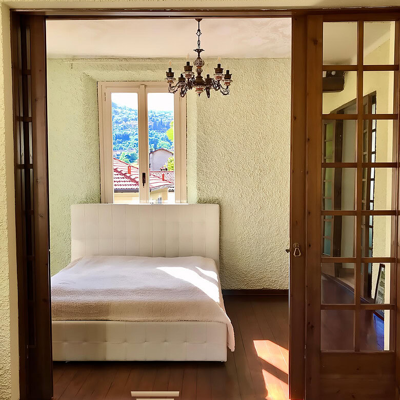 Уютная спальня с видом на природу, светлые стены и деревянный пол, просторное окно и стильная люстра, современная мебель и природное освещение, идеально подходит для комфорта и отдыха.