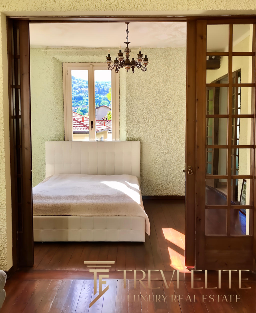 Уютная спальня с видом на природу, светлые стены и деревянный пол, просторное окно и стильная люстра, современная мебель и природное освещение, идеально подходит для комфорта и отдыха.