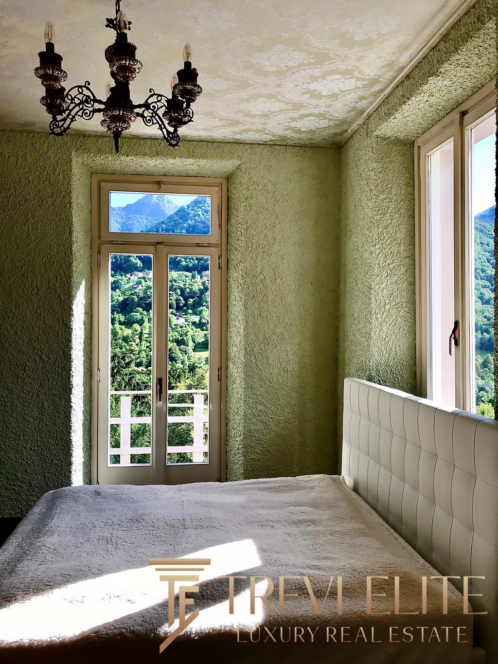 уютная спальня с панорамными окнами и видом на горы, светлые стены с текстурой, стильная люстра и мягкое постельное белье, создающее уют и комфорт в премиальной недвижимости Треви Элит.