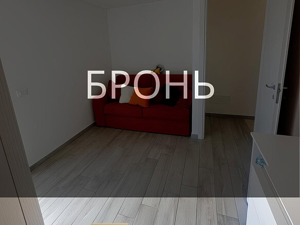 ОПИСАНИЕ ИЗОБРАЖЕНИЯ: Внутренний вид уютной комнаты с светлой деревянной отделкой пола, белыми стенами и простой потолочной конструкцией. На стене расположен светильник без абажура, в углу стоит красный диван с подушками, за дверью виднеется другой небольшой сухой коридор. Логотип агентства недвижимости "Треви Элит" и название "БРОНЬ" наложены на изображение.