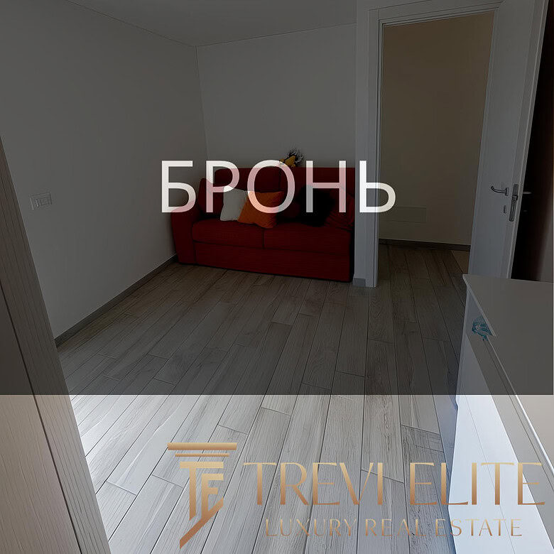 ОПИСАНИЕ ИЗОБРАЖЕНИЯ: Внутренний вид уютной комнаты с светлой деревянной отделкой пола, белыми стенами и простой потолочной конструкцией. На стене расположен светильник без абажура, в углу стоит красный диван с подушками, за дверью виднеется другой небольшой сухой коридор. Логотип агентства недвижимости "Треви Элит" и название "БРОНЬ" наложены на изображение.
