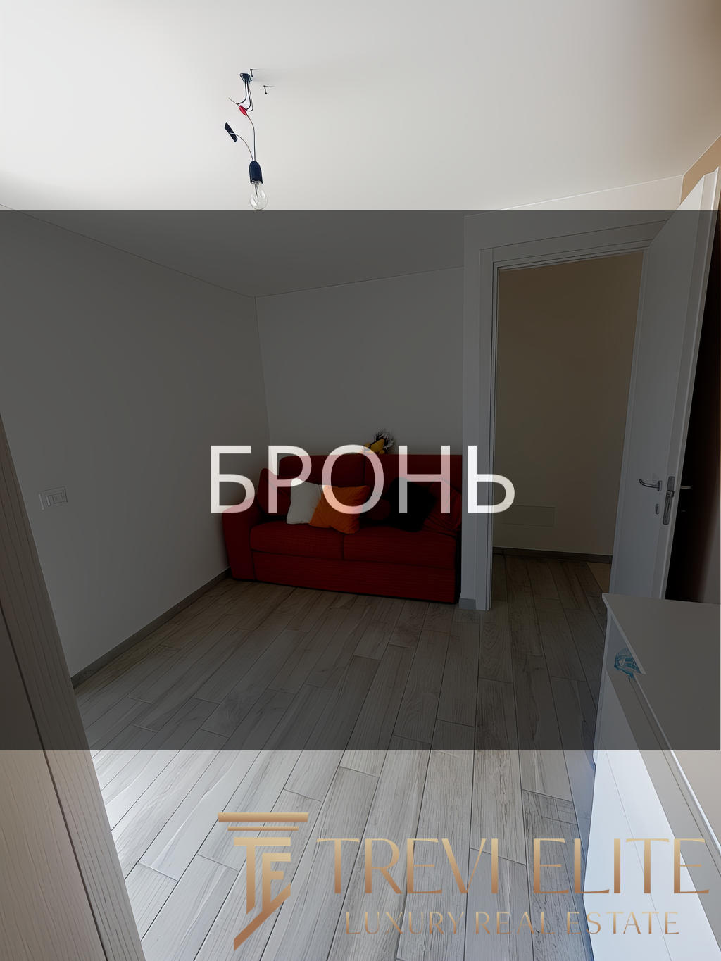 ОПИСАНИЕ ИЗОБРАЖЕНИЯ: Внутренний вид уютной комнаты с светлой деревянной отделкой пола, белыми стенами и простой потолочной конструкцией. На стене расположен светильник без абажура, в углу стоит красный диван с подушками, за дверью виднеется другой небольшой сухой коридор. Логотип агентства недвижимости "Треви Элит" и название "БРОНЬ" наложены на изображение.
