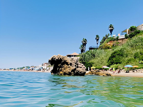 Роскошный морской пейзаж с пляжем, скалами и зеленью у побережья, солнечная погода, голубое небо, курортная атмосфера, строительство на склоне горы, пальмы, пляжные зонты, спокойная вода, витриальные виды элитной недвижимости.