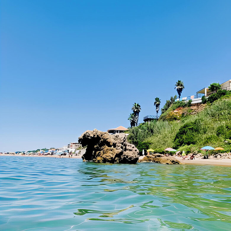 Роскошный морской пейзаж с пляжем, скалами и зеленью у побережья, солнечная погода, голубое небо, курортная атмосфера, строительство на склоне горы, пальмы, пляжные зонты, спокойная вода, витриальные виды элитной недвижимости.