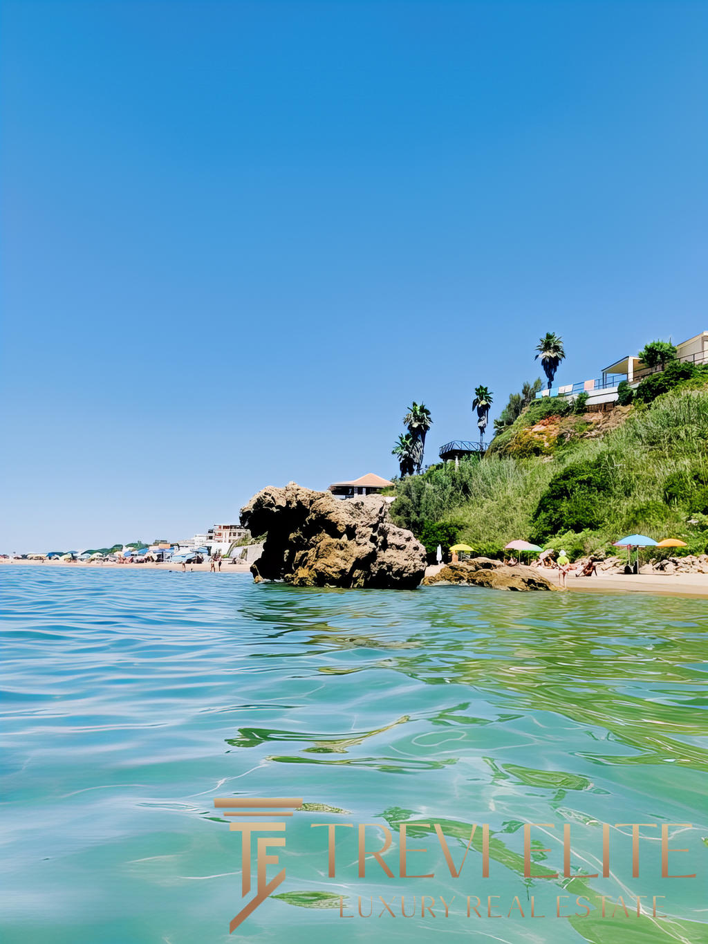 Роскошный морской пейзаж с пляжем, скалами и зеленью у побережья, солнечная погода, голубое небо, курортная атмосфера, строительство на склоне горы, пальмы, пляжные зонты, спокойная вода, витриальные виды элитной недвижимости.