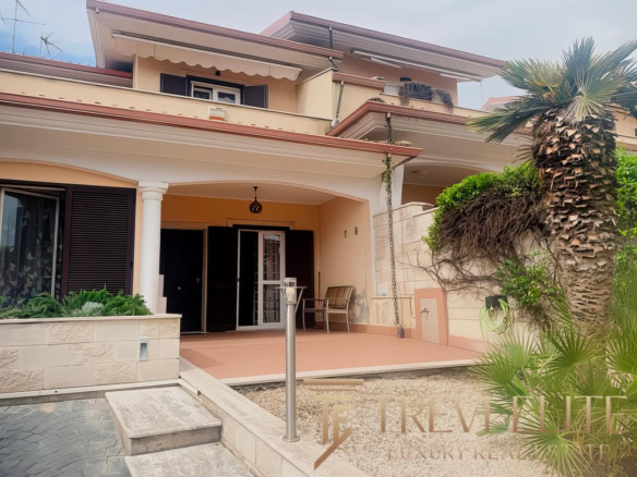 Открытая терраса современного жилого дома с пальмой и зеленью, вход в дом с дверью и зоной отдыха, уличное освещение и уютная атмосфера, стильный дизайн загородного жилья, агентство недвижимости Треви Элит.