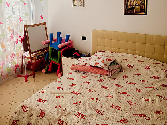 Качественный интерьер светлой спальни с двухспальной кроватью, украшенной постельным бельем с красными узорами, детские игрушки и зеркало у окна, стильная и уютная обстановка.