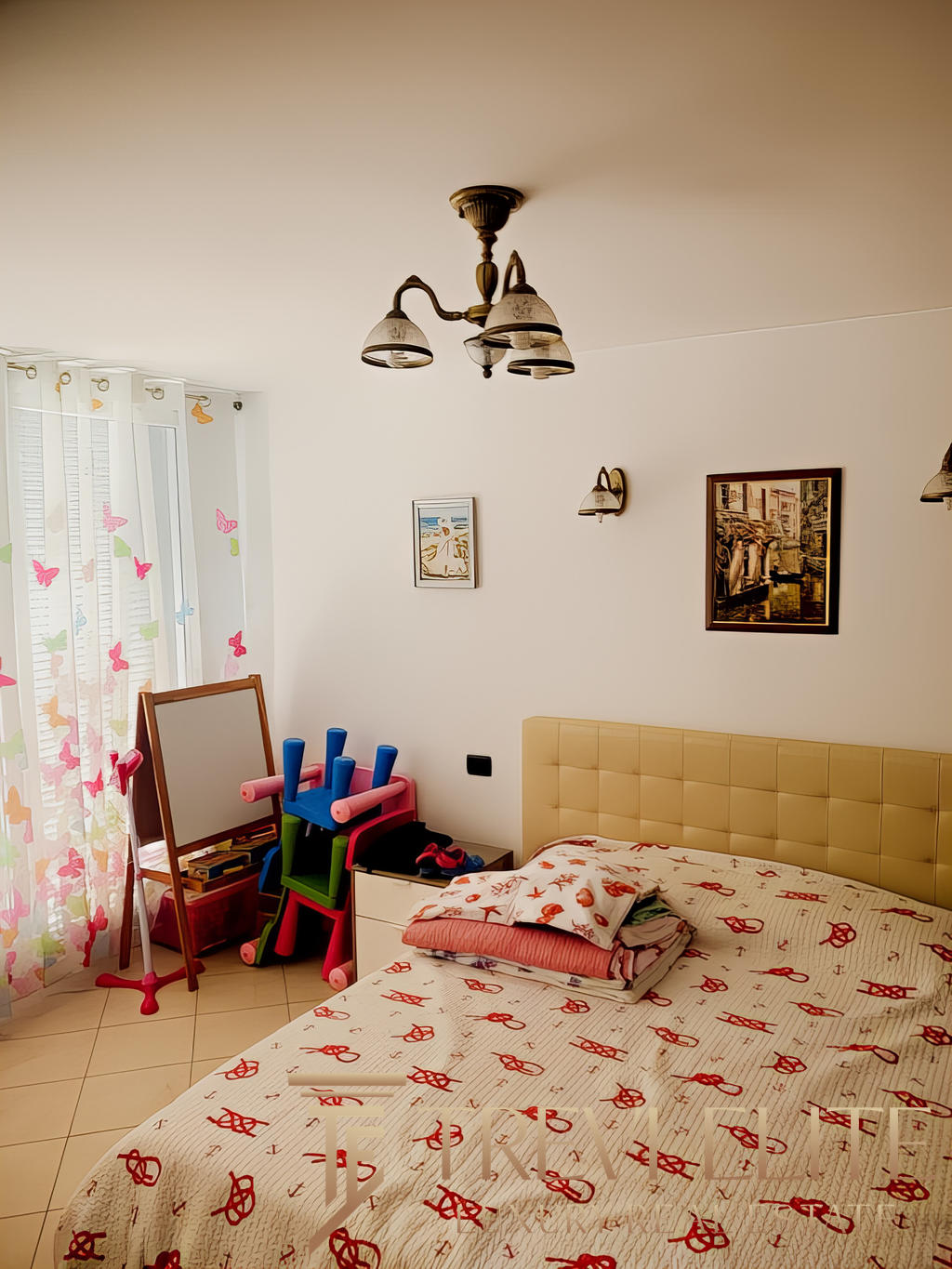 Качественный интерьер светлой спальни с двухспальной кроватью, украшенной постельным бельем с красными узорами, детские игрушки и зеркало у окна, стильная и уютная обстановка.