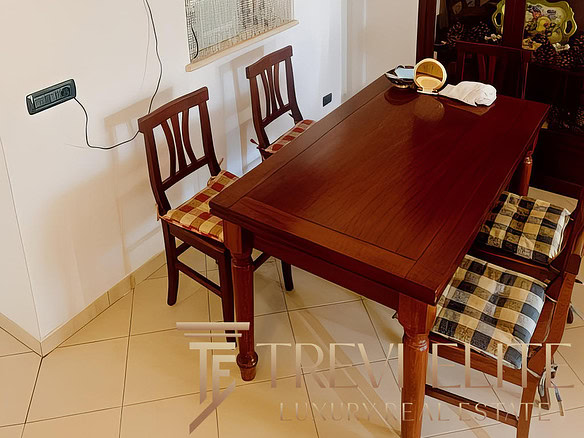 Столовая зона в уютной квартире с деревянным столом и стульями, стеклянный шкаф с посудой и декоративными предметами, мягкое освещение, украшения на стенах и полке, создающие теплая атмосферу.