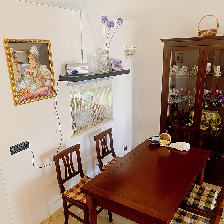 Столовая зона в уютной квартире с деревянным столом и стульями, стеклянный шкаф с посудой и декоративными предметами, мягкое освещение, украшения на стенах и полке, создающие теплая атмосферу.