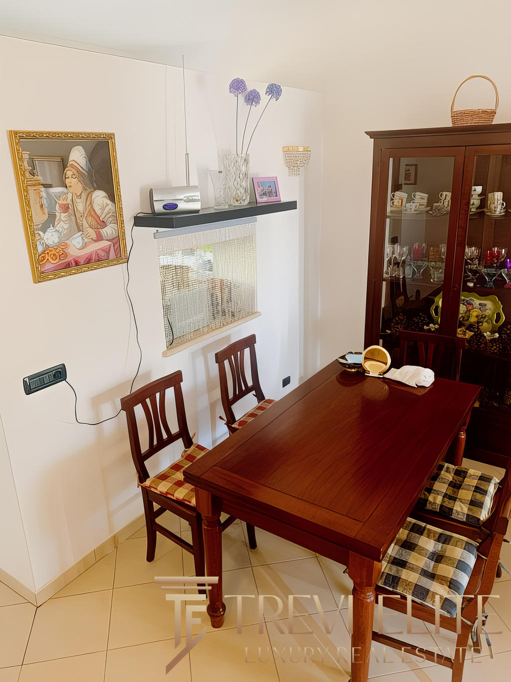 Столовая зона в уютной квартире с деревянным столом и стульями, стеклянный шкаф с посудой и декоративными предметами, мягкое освещение, украшения на стенах и полке, создающие теплая атмосферу.