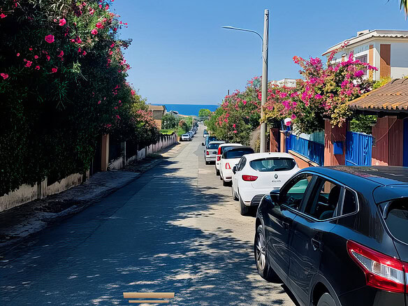 Булыжная улица с парками и цветущими деревьями рядом с морем, перспективы на побережье, автомобили на дороге, солнечный день, спокойная романтичная атмосфера.