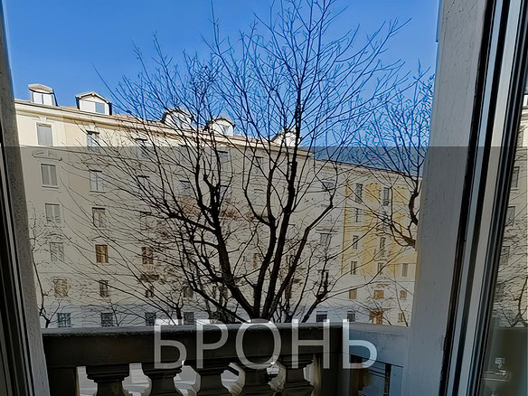 Вид из окна на зимний двор с деревьями и многоэтажными зданиями, солнечный день, красивый городской пейзаж, балкон с классической балюстрадой, просторное помещение, светлый интерьер.