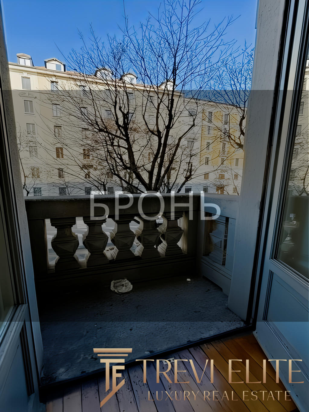 Вид из окна на зимний двор с деревьями и многоэтажными зданиями, солнечный день, красивый городской пейзаж, балкон с классической балюстрадой, просторное помещение, светлый интерьер.