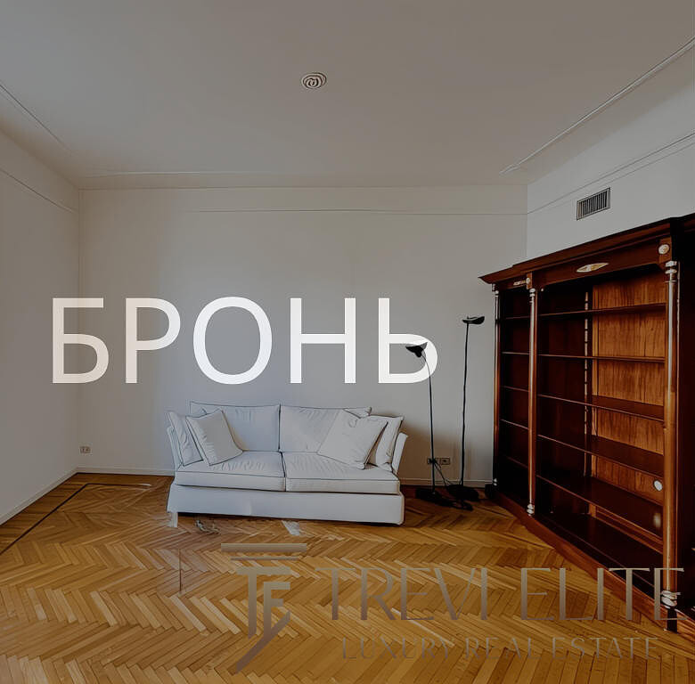 Современная уютная гостиная с деревянным полом и большой книгой полкой, светлым диваном, окнами и минималистичным дизайном.