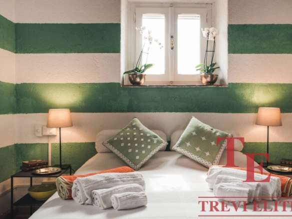 Яркая спальня с зелеными полосатыми стенами, уютной кроватью с декоративными подушками и камерой с двумя лампами, создающими тёплую атмосферу.