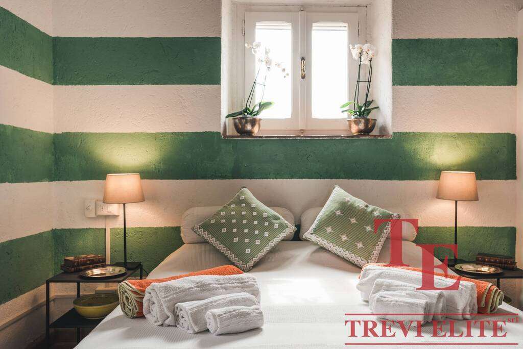 Яркая спальня с зелеными полосатыми стенами, уютной кроватью с декоративными подушками и камерой с двумя лампами, создающими тёплую атмосферу.