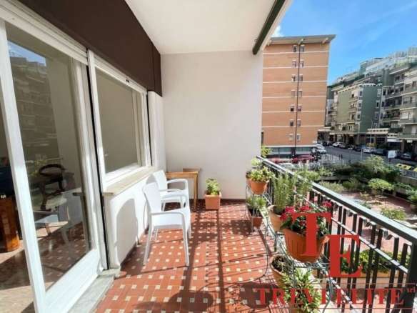 Тёплый просторный балкон с цветущими растениями и удобной мебелью, идеально подходящий для отдыха и наслаждения видом на жилые дома и городскую застройку в центре города.