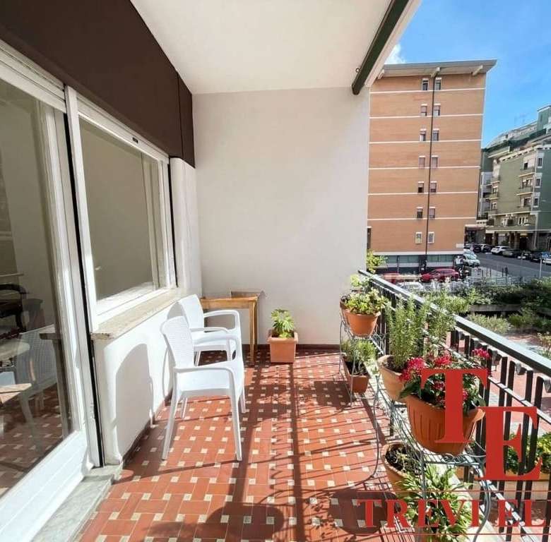 Тёплый просторный балкон с цветущими растениями и удобной мебелью, идеально подходящий для отдыха и наслаждения видом на жилые дома и городскую застройку в центре города.