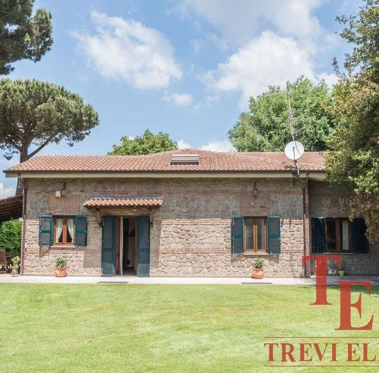 Испанский загородный дом с каменной облицовкой, окруженный зелёным газоном и высокими деревьями, под ясным небом, сочетающий элегантность и уединение, эксклюзивное предложение от Треви Элит.