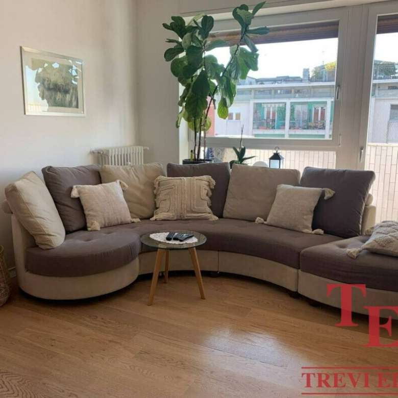 Стильная гостиная с большими окнами, уютным угловым диваном и зелёными растениями, создающими атмосферу комфорта и роскоши в интерьере современного элитного жилья.