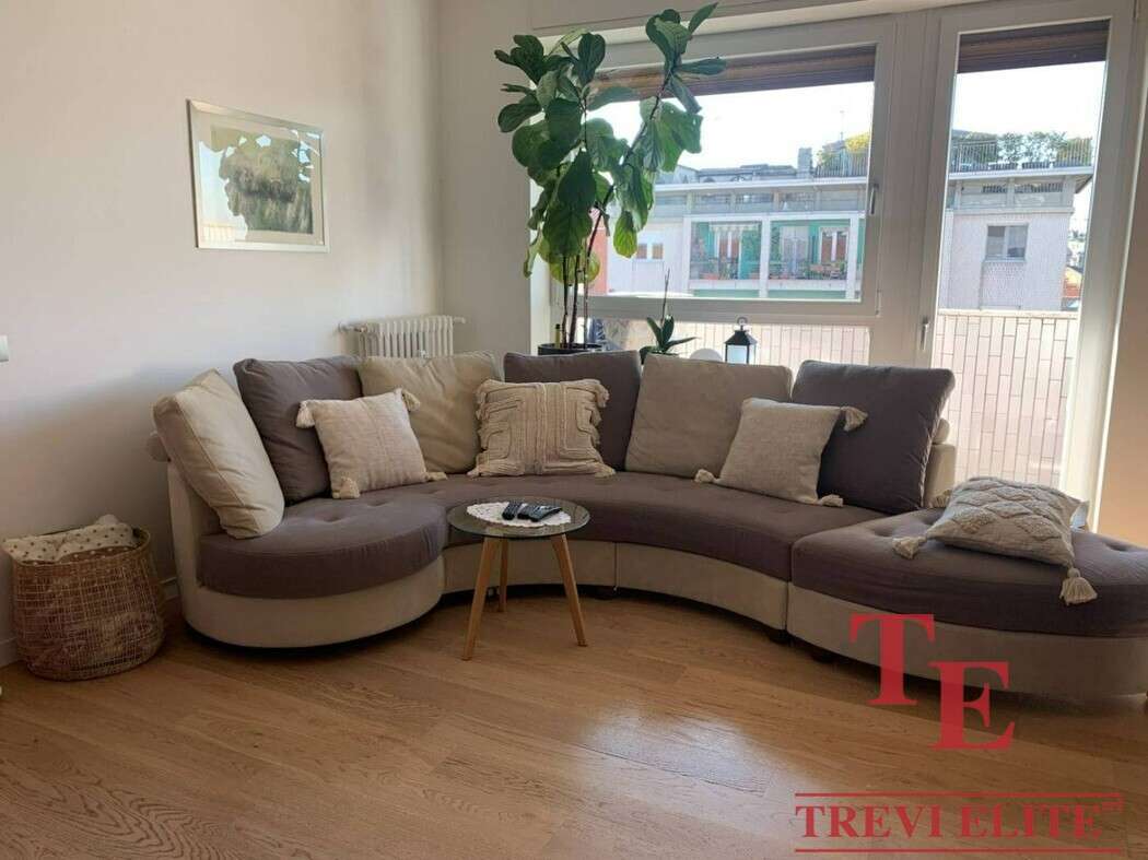 Стильная гостиная с большими окнами, уютным угловым диваном и зелёными растениями, создающими атмосферу комфорта и роскоши в интерьере современного элитного жилья.