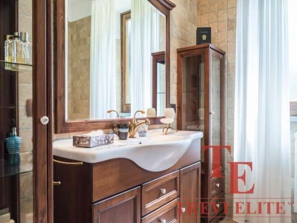 Элегантная ванная комната с деревянной мебелью, большой раковиной и зеркалом, уютным освещением и солнечным естественным светом через большие окна с белыми шторами.