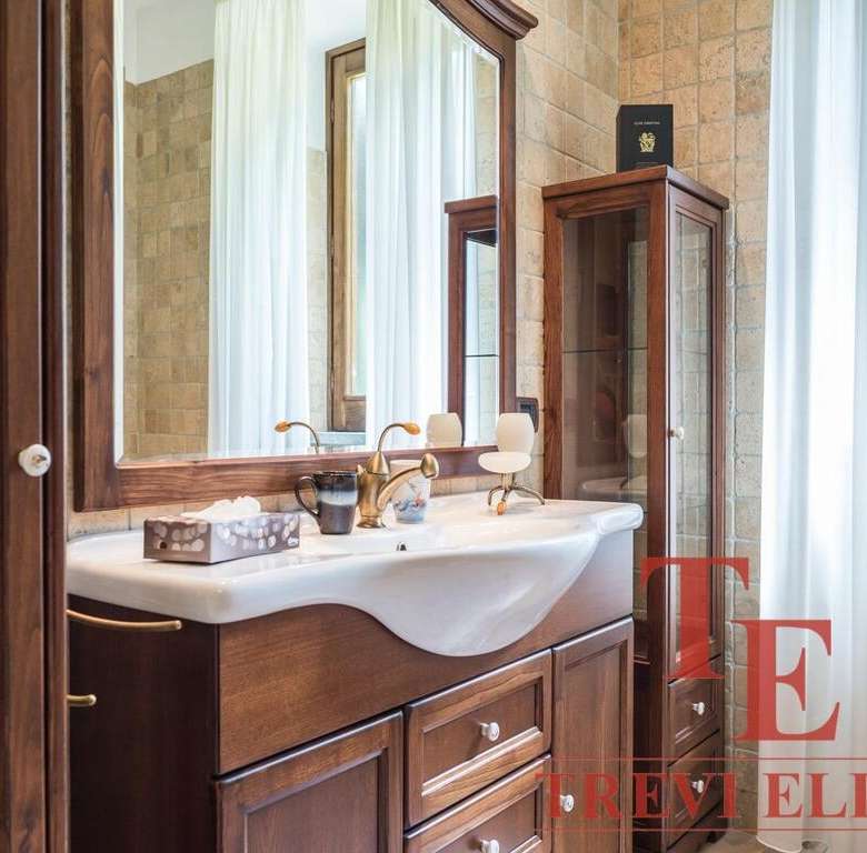 Элегантная ванная комната с деревянной мебелью, большой раковиной и зеркалом, уютным освещением и солнечным естественным светом через большие окна с белыми шторами.