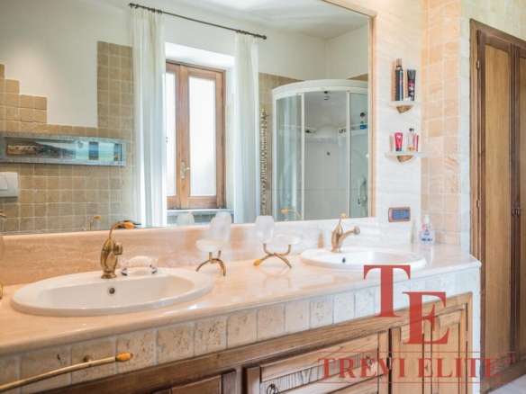 Роскошная ванная комната с двумя раковинами, деревянными шкафчиками, большой зеркальной панелью и современным душем. Светлые тона и природное освещение создают уютную атмосферу.