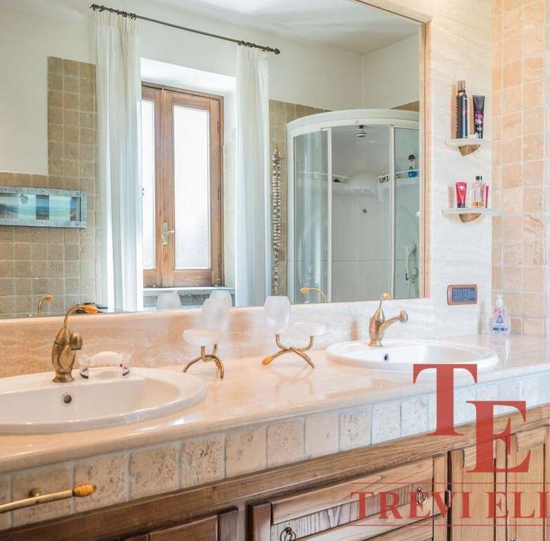 Роскошная ванная комната с двумя раковинами, деревянными шкафчиками, большой зеркальной панелью и современным душем. Светлые тона и природное освещение создают уютную атмосферу.