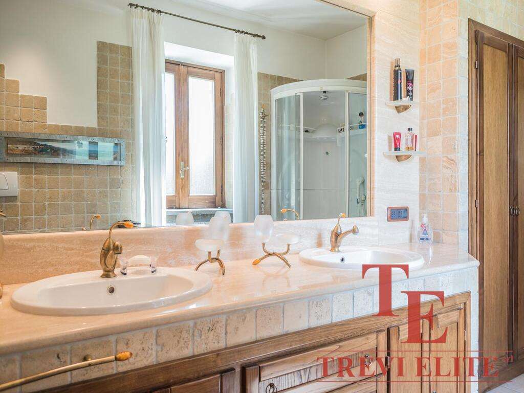 Роскошная ванная комната с двумя раковинами, деревянными шкафчиками, большой зеркальной панелью и современным душем. Светлые тона и природное освещение создают уютную атмосферу.