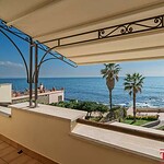 Вид на море с просторного балкона с тентом, окруженного зеленью и пальмами, солнечный день, яхты на горизонте, роскошное жилье в престижном районе.