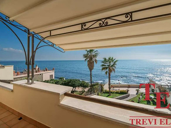 Вид на море с просторного балкона с тентом, окруженного зеленью и пальмами, солнечный день, яхты на горизонте, роскошное жилье в престижном районе.